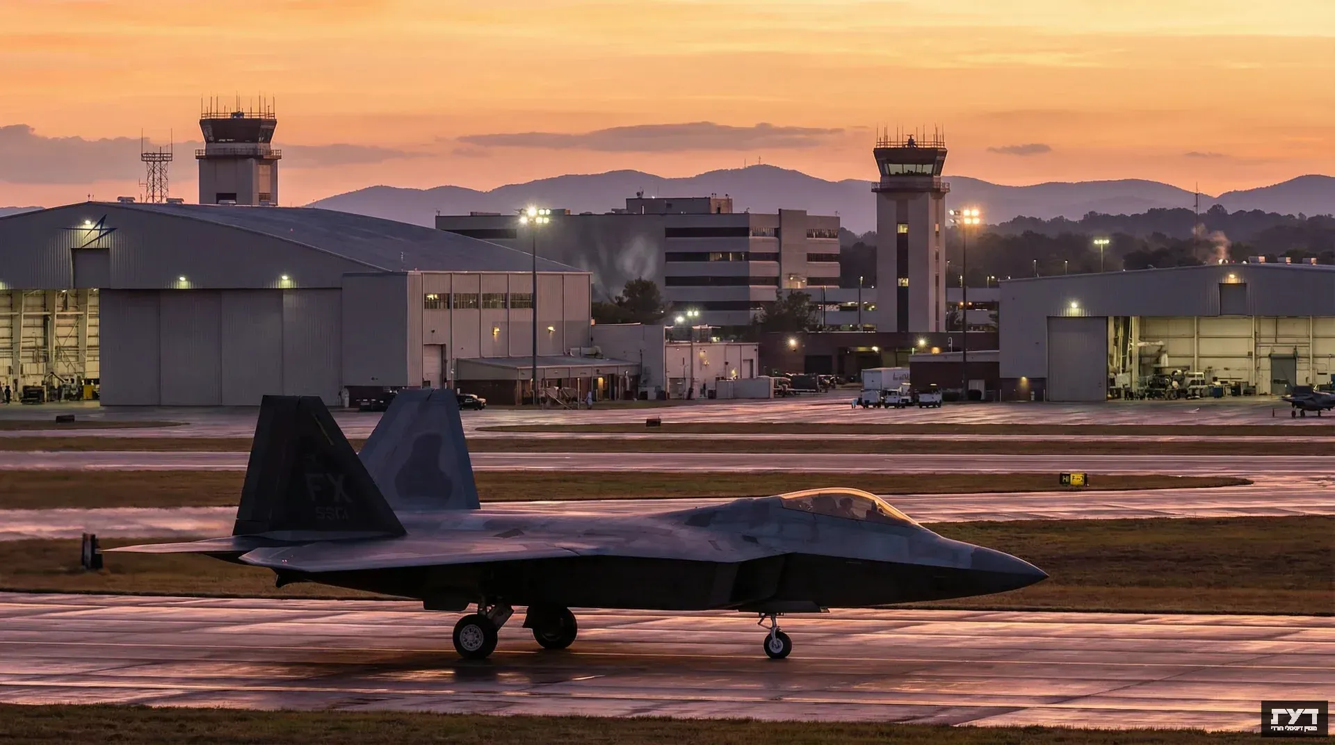 מטוס קרב F-22 ראפטור על המסלול במתקן לוקהיד מרטין בשעת דמדומים, מגדלי פיקוח והאנגרים ברקע