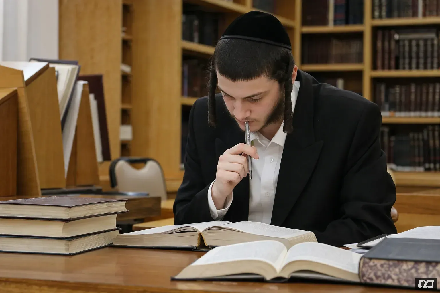 סטודנט חרדי יושב בחדר לימוד מוקף ספרים פתוחים, מחזיק עט ומרוכז בלמידה, ברקע ארונות ספרים ותאורה חמה