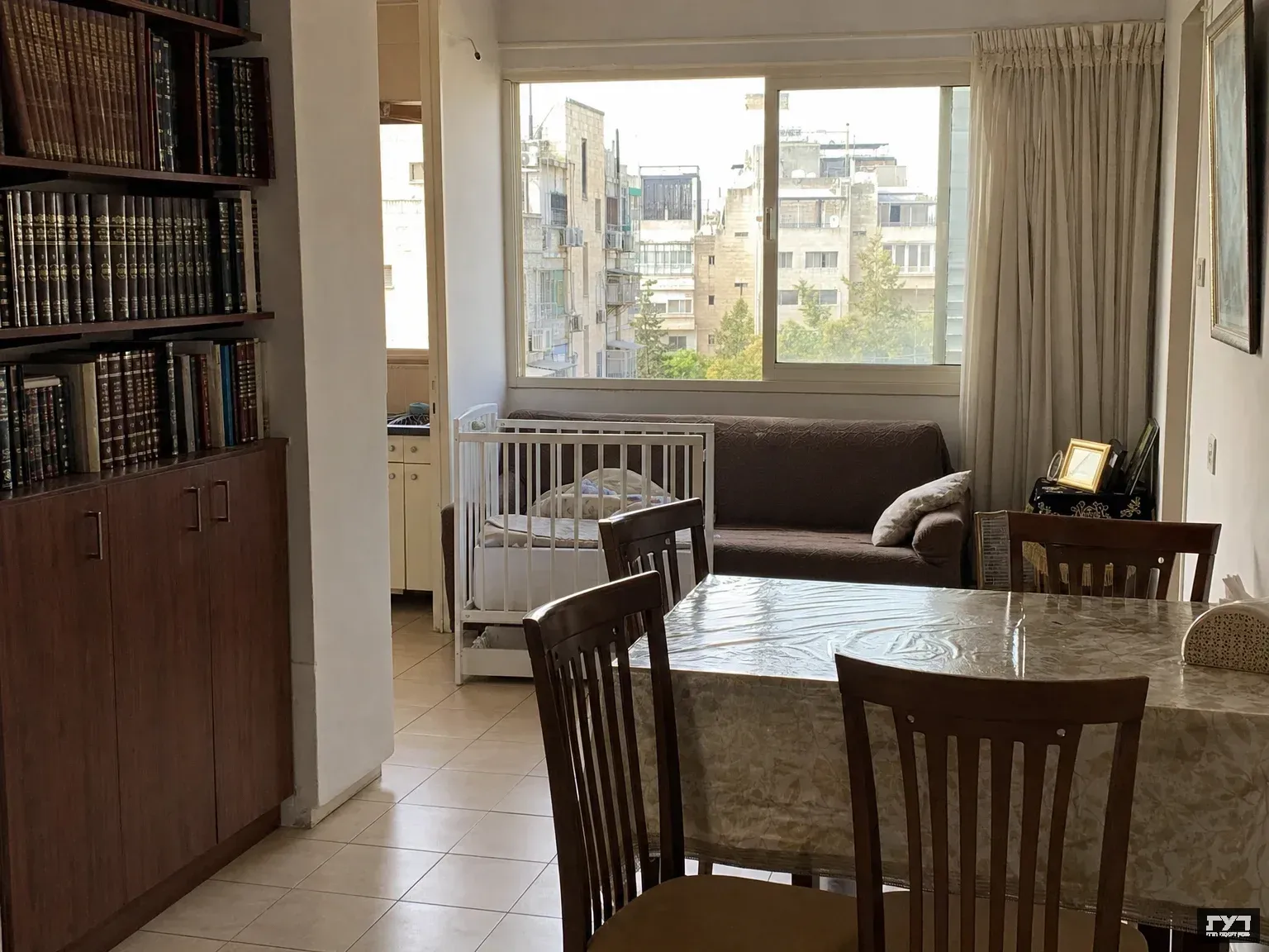 פנים דירה חרדית צנועה: ספריית ספרי קודש, שולחן אוכל מכוסה ניילון, מיטת תינוק וחלון עם נוף עירוני