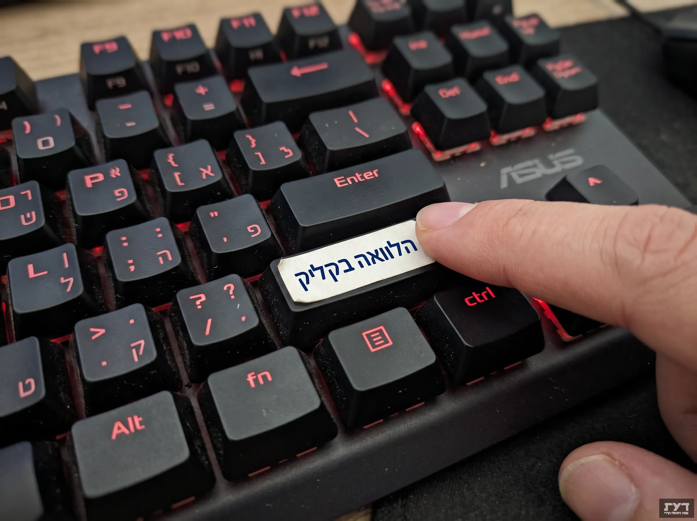 בתמונה נראית מקלדת גיימינג מכנית שחורה של מותג ASUS עם תאורה אחורית אדומה. אצבע של יד אדם לוחצת על מקש ה-Shift הימני, שהוחלף במקש לבן מותאם אישית. על המקש הלבן מופיע הכיתוב בעברית "הלוואה בקליק" בצבע כחול.